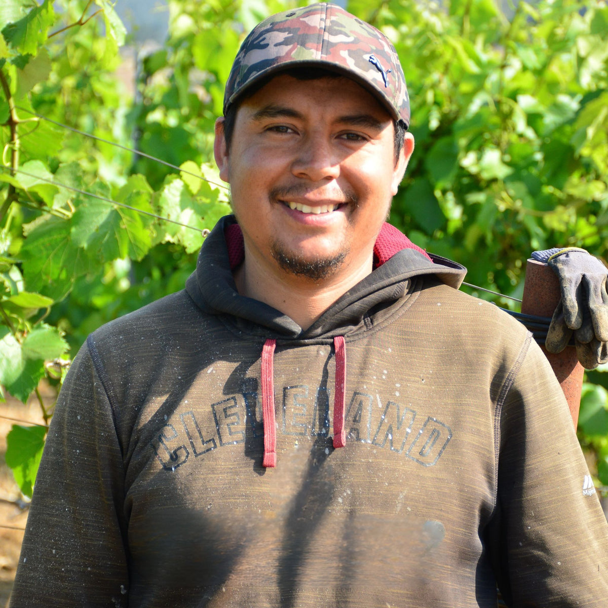 A smiling man wearing a camouflage baseball cap, a dark brown Cleveland hoodie, and a work glove, standing outdoors in front of a green vineyard background.