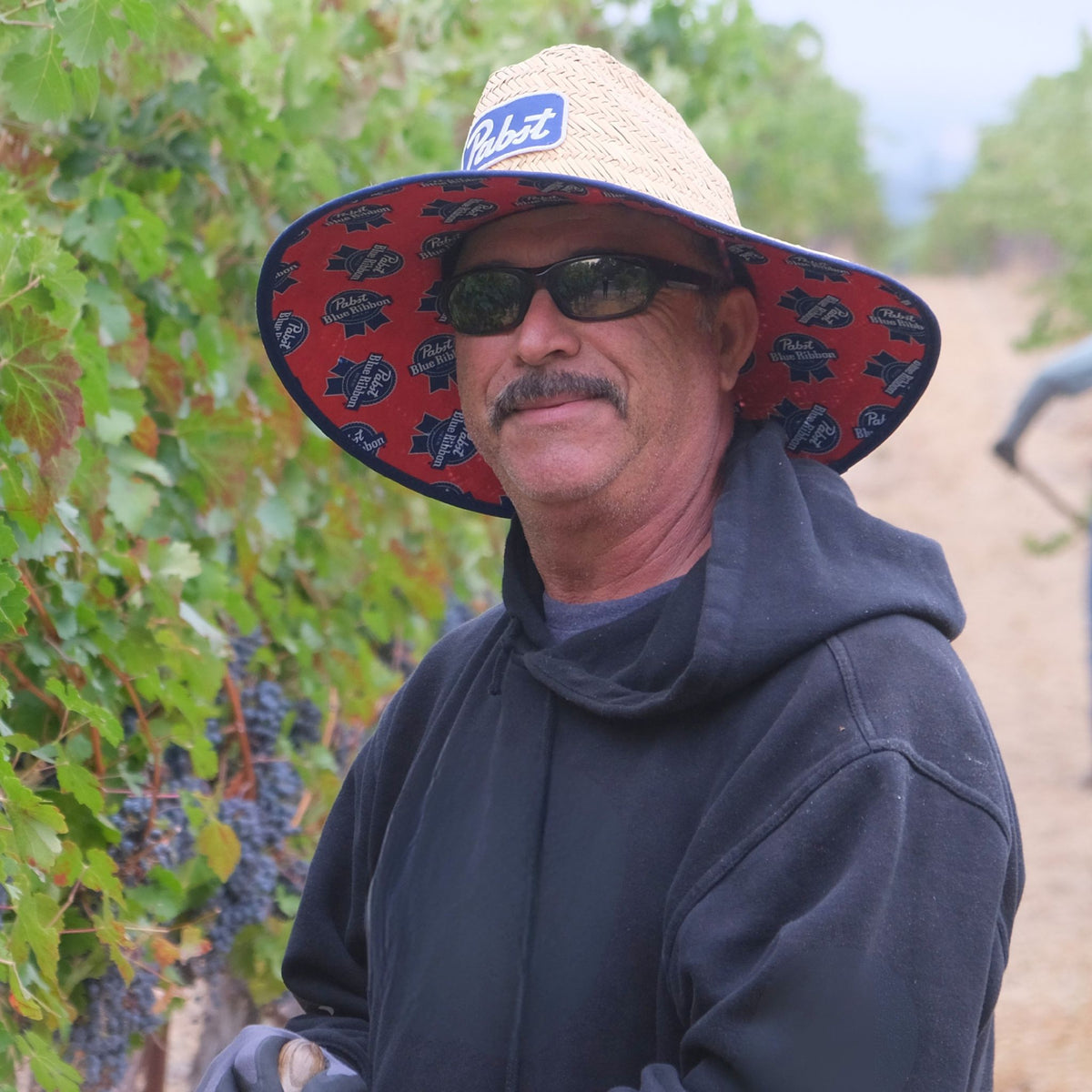 A man wearing a wide-brimmed straw hat with a red patterned brim and a blue brand patch, along with dark sunglasses and a dark hoodie, standing in a vineyard.