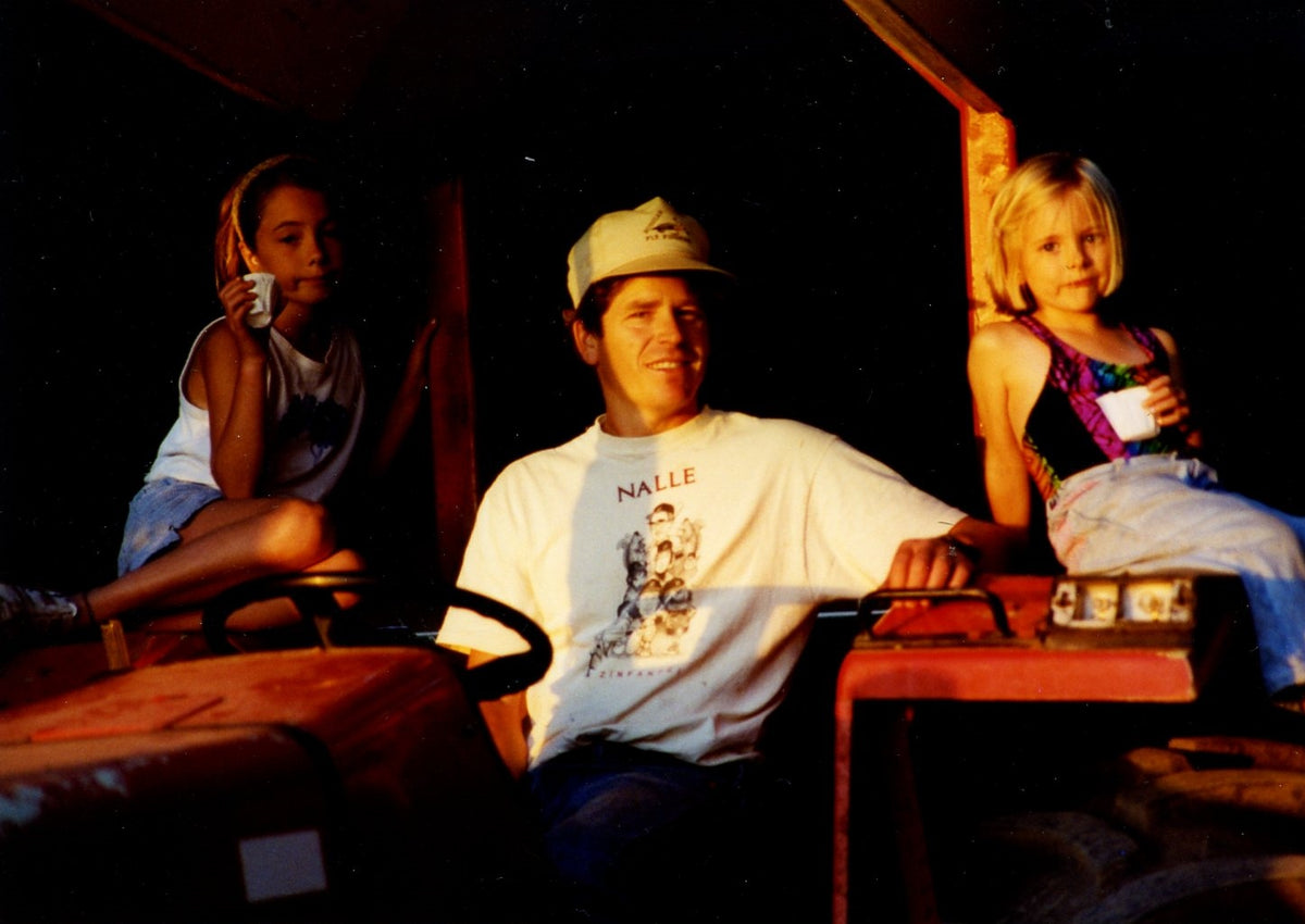 A man wearing a white graphic t-shirt and a baseball cap sits in the driver's seat of a vehicle, with a young girl on his right and another girl on his left, photographed in low-light conditions.