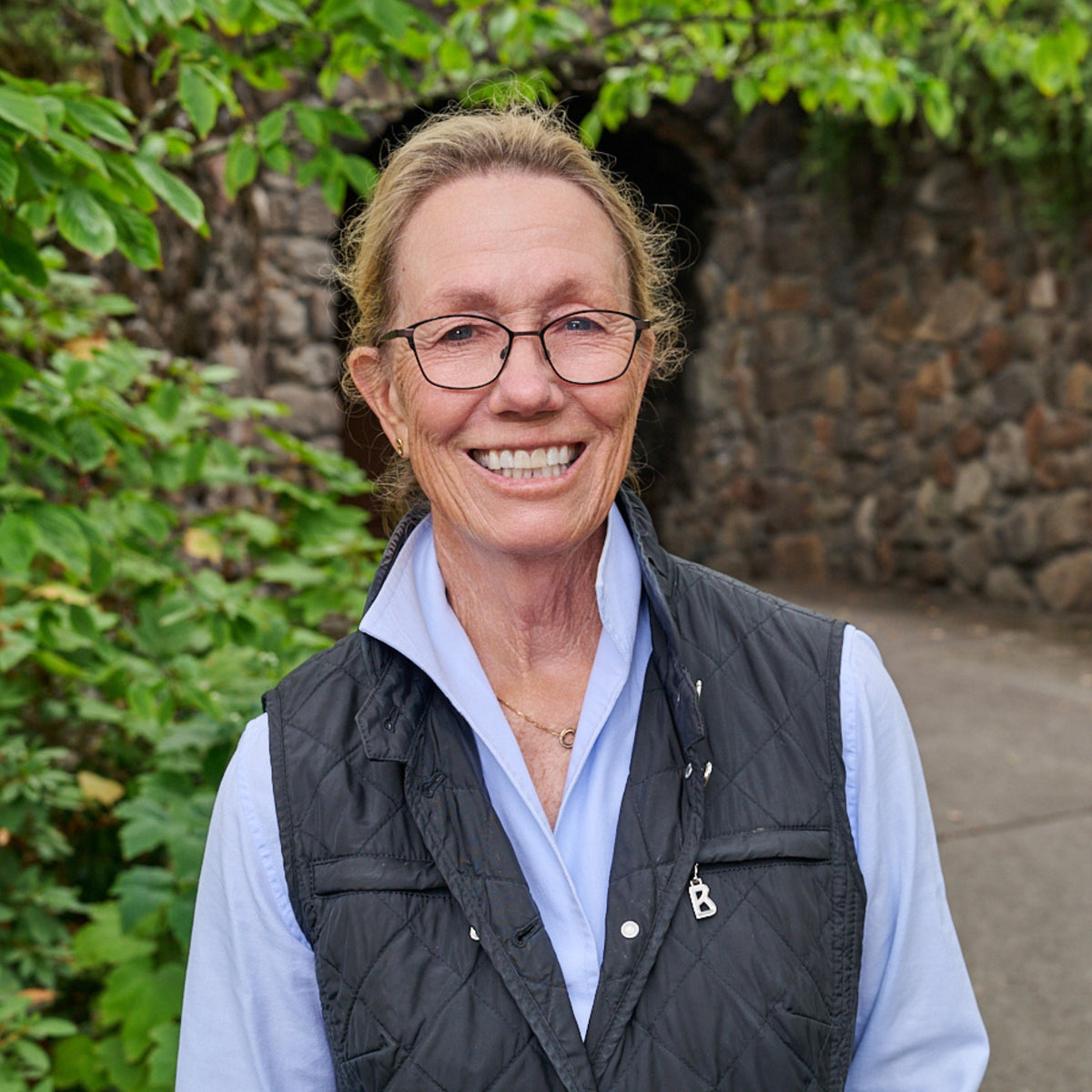 Sarah Hafner smiling with glasses, wearing a black quilted vest over a light blue button-up shirt, standing outdoors in front of a stone wall and green foliage.
