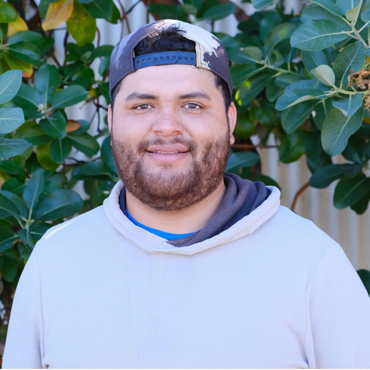 A man wearing a light gray hoodie, a blue t-shirt, a dark gray and white patterned baseball cap, and a dark gray neck warmer, standing outdoors in front of green foliage.